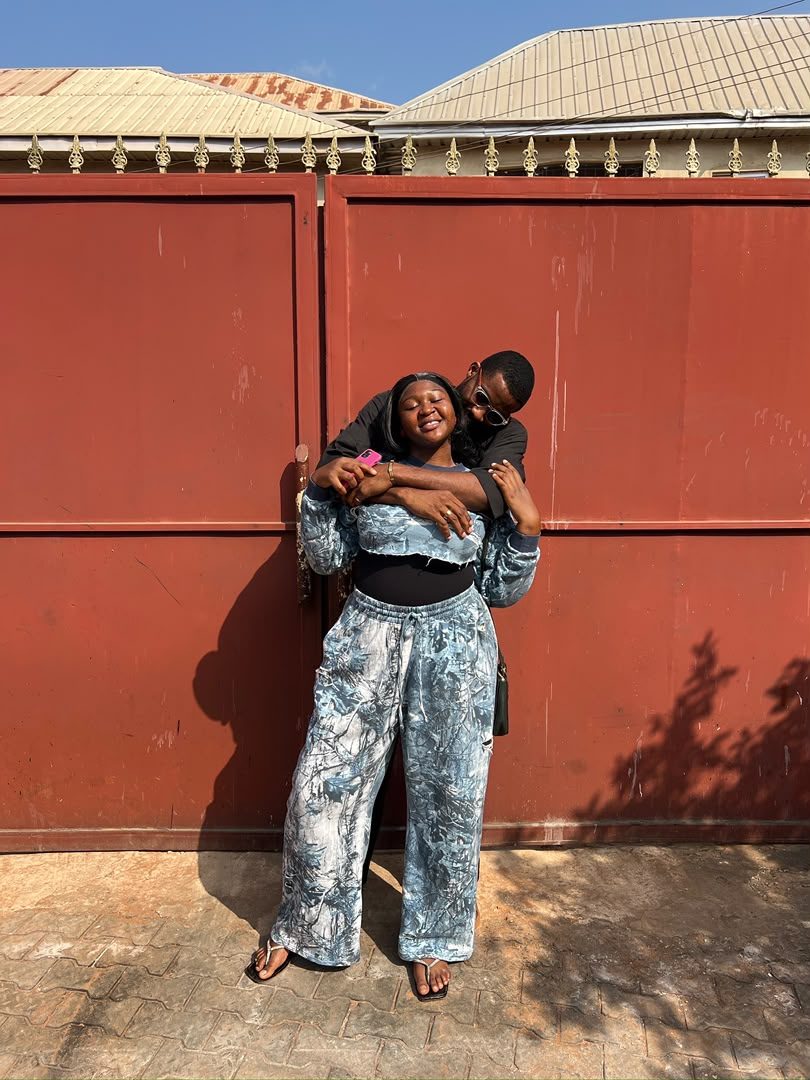 Chukwuma embracing Nnenna from behind in front of a red gate
